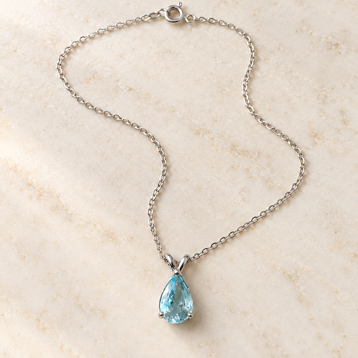 Sky-blue pear-cut pendant on a fine silver-tone chain – laid in a curve on an ivory stone surface