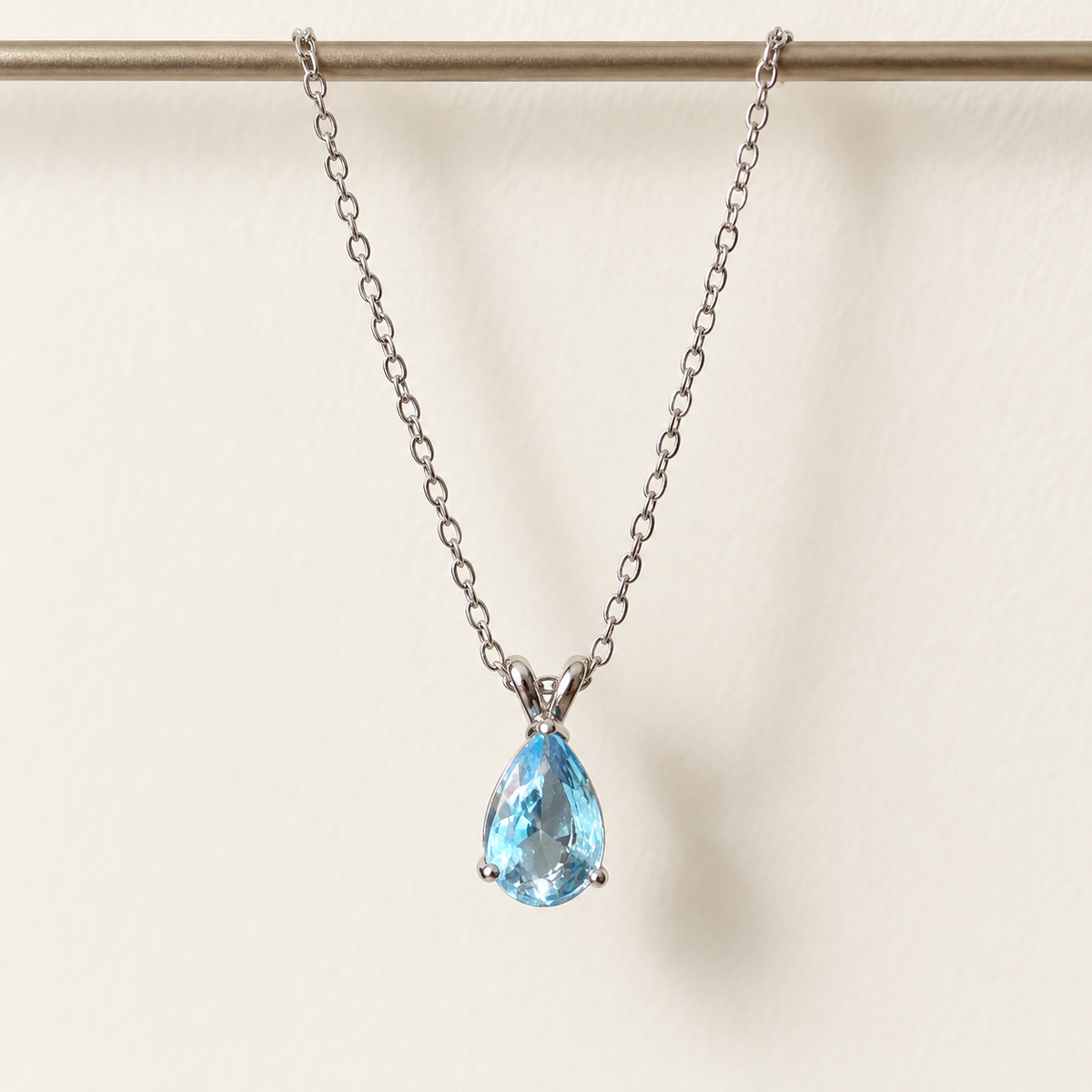 Sky-blue pear-cut pendant on a fine silver-tone chain – hanging straight against an ivory wall